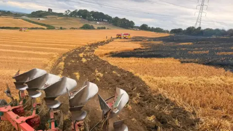 Penshaw Padel Agricultural machinery with metal drill-bits in the left hand corner of the photo. It is ploughing harvested wheat fields beside a large area of blackened, burned fields. There is a fire engine positioned in the distance under an overcast sky.
