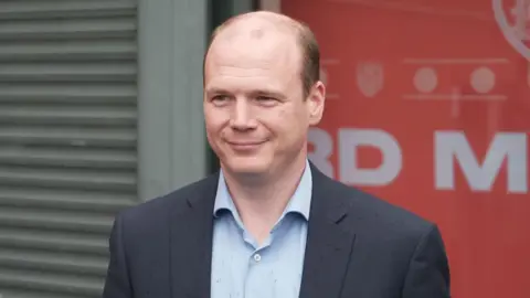 Lyons is wearing a grey blazer and a light blue shirt. He has brown hair around the side of his head and balding on top. He has a slight smile. 
