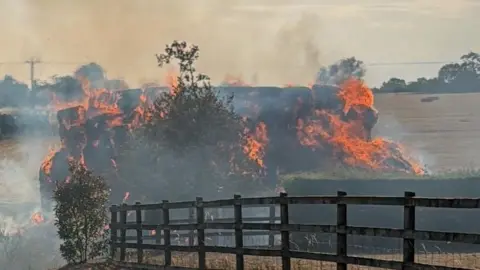 Bob Brace A large haystack on fire in a field by the side of a road, close to a fence and hedging. Orange flames and smoke are coming off the stacked hay and there are fields, hedgerows and trees in the background. 