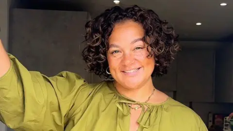 Grace, who has short brown curly hair, smiles while wearing a green top