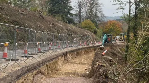 Work to the embankment along the A36. A construction worker is using a digger to clear trees and bushes. Metal fencing can be seen along the roadside with orange traffic cones.