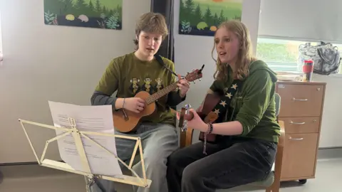 Harrie Wilson and Cora Tarling sat with a guitar and ukulele playing in a hospital ward.
