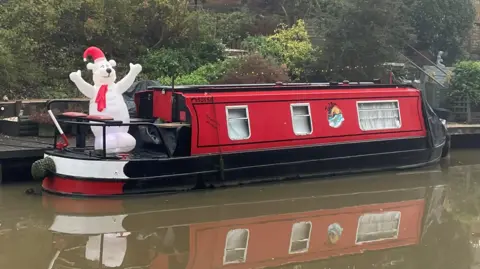 John Sadler A Christmas polar bear with a red scarf and hat is standing on a red boat on the canal.