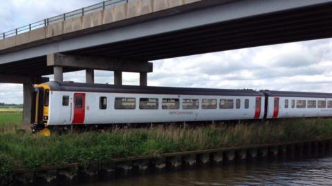 A Greater Anglia train passes beneath a concrete bridge beneath a cloudy sky. There are fields in the distance and a river and green grass in the foreground