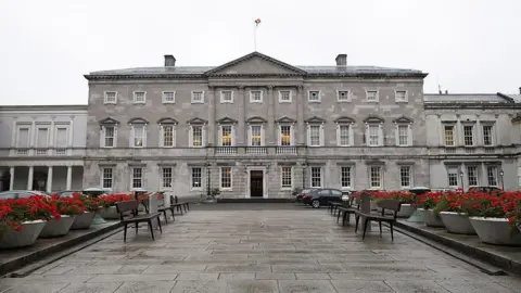 A general view of Leinster House which houses the Seanad chamber, also known as the upper house of the Irish parliament, is pictured in Dublin, Ireland, in 2013. It is a large grey Georgian, Palladian style building with the Irish flag on top. A row of red flowers in pots and seating benches line a walkway leading up to it.
