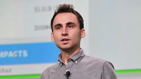 Nikita Bier, wearing a grey, button-up shirt with a microphone attached, speaks on-stage at TechCrunch Disrupt in 2013.