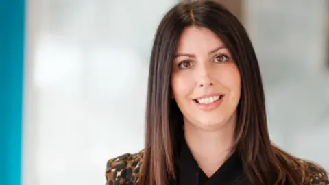 Hudgell Solicitors Hayley Collinson has long dark hair. She is wearing a dark brown and black top and is looking at the camera with a serious smile. 