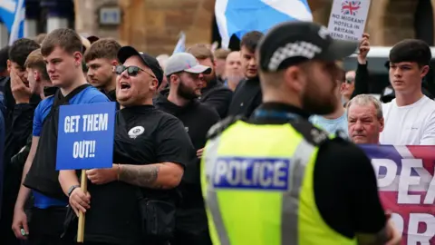 A group of men in dark clothing, one holds a blue sign which says "GET THEM OUT". A police officer faces the crowd and his high vis vest can be seen in the foreground 