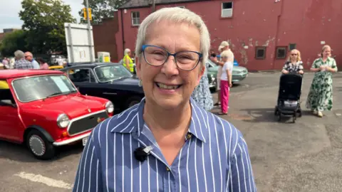 A woman in a stripy blouse, with short grey hair and glasses smiles widely with classic cars and people milling around behind her