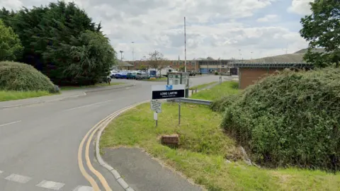 A road junction into Long Lartin Prison with a black and white Long Lartin sign in the grass near a bush. A car park can be seen in the background along with multiple brick buildings.