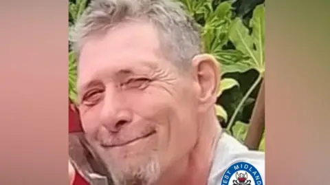 A photograph of Stephen Peek before he died. He smiles at the camera with greenery behind him. He has short greying hair, moustache and beard.
