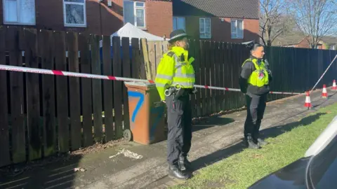 Two police officers stood in front of a police tape cordon, with a wooden fence and housing behind