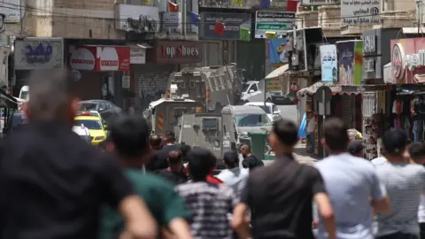 Palestinian men are standing on a street in Nablus on 27 May. Israeli tanks are in the background while shops are at the side