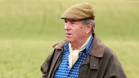 Charles Collingwood wearing a coat, chequered blue and white shirt, white neckerchief and flat cap stood in a field being used for crops