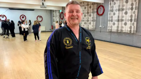 John is wearing a navy/black Taekwondo suit. He has a beard and short grey/brown hair. Women in the class can be seen behind him
