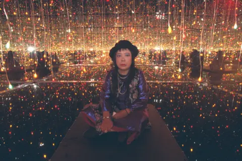 Getty Images Yayoi Kusama in 2001 seated on the floor of one of her installation rooms, surrounded by hundreds of small coloured lights and mirrors