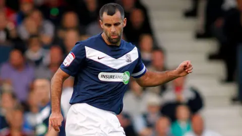 Chris Barker appears to be stretching to kick the ball during a match. He is wearing a blue Southend United strip and is wearing the captain's armband. There is a crowd sitting in the stands in the background.