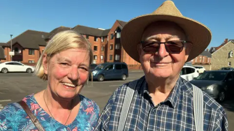 A couple stand outside of a council drop-in session. The woman on the left has blonde hair cut into a short bob, and is wearing a blue patterned blouse. The man on the right is wearing a blue checked shirt, grey suspenders, black sunglasses and a straw brimmed hat.