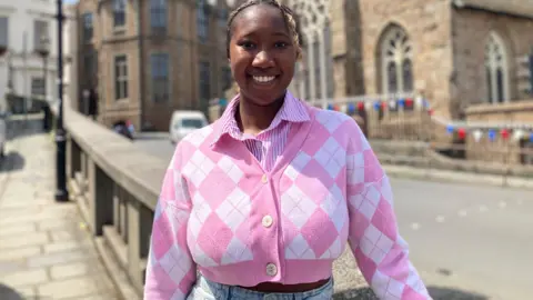 A woman in a pink patterned top with a number of buildings behind her