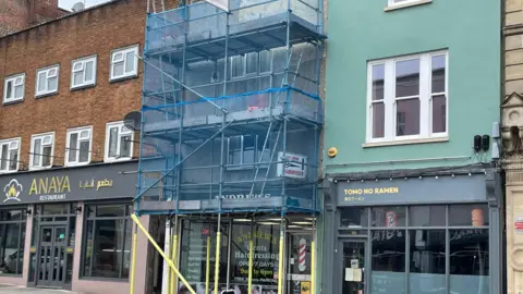 A high street where one shop is covered in scaffolding. Blue netting covers the scaffolding. Signage for Andrew's Gentleman Hairdresser is just visible.
