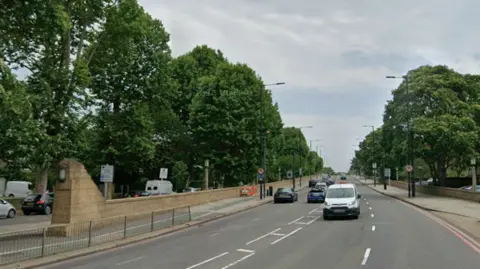 Google Street View image of a wide, tree‑lined urban road with light traffic and pavements on both sides.