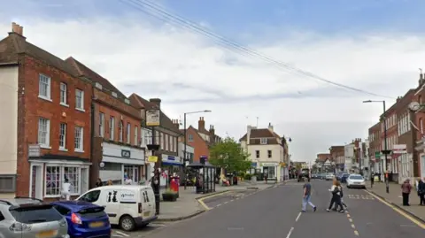 Witham town centre. There is a road with people crossing it and cars parked on either side. There are also shops that line the road.