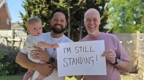 Two men smile at the camera while holding a baby and a piece of white paper saying 'I'm still standing!'