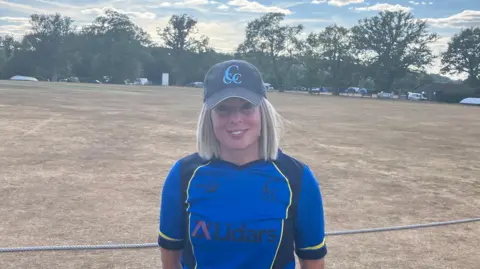 En la fotografía, Lucy Merredy aparece al borde del campo de críquet. Lleva una camiseta azul y una gorra azul marino. Tiene el pelo rubio hasta los hombros.