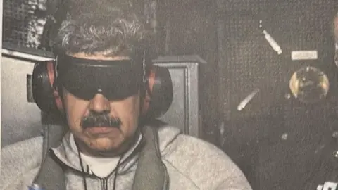 Donald Trump Nicolas Maduro shown after his arrest, wearing headphones and a blindfold, grey zipped jumper