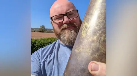 Ross Miller, who has a beard and wears glasses, is holding a large metal cylinder which is a 1940s bombshell.