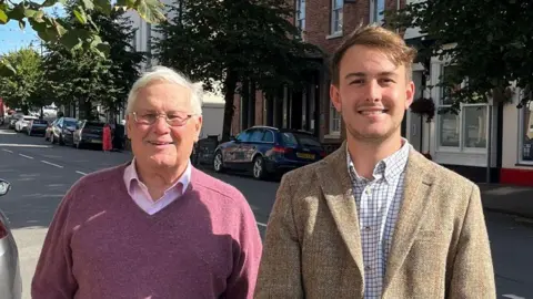 Gareth Morgan (left) stands next to Glyn Preston (right) in a street. They both smile at the camera in the sunshine. Mr Morgan wears a purple v-neck jumper and pink shirt, he has silver short hair and wears thin rimmed sunglasses. Mr Preston wears a tweed suit jacket and a white checked shirt and has light brown hair. 