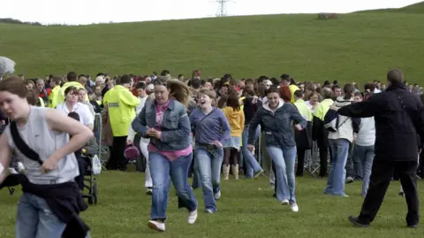 Several young women run into a field after being let through a security gate behind which a crowd is gathered. A man in black is gesturing at them to slow down.