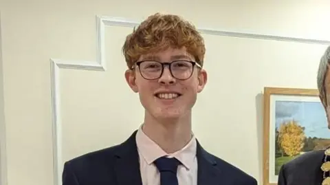 Amanda Morgan A teenage boy with ginger hair and black glasses. He is wearing a navy suit.