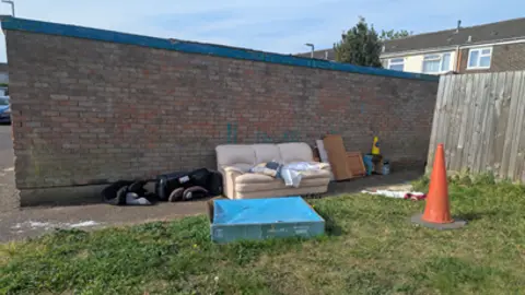 RSPCA Rubbish including a cream-coloured sofa, wooden furniture, orange traffic cone and a child's car seat abandoned next to a brick wall on a housing estate. 