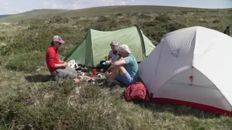 Three people sit next to two tents on open moorland.