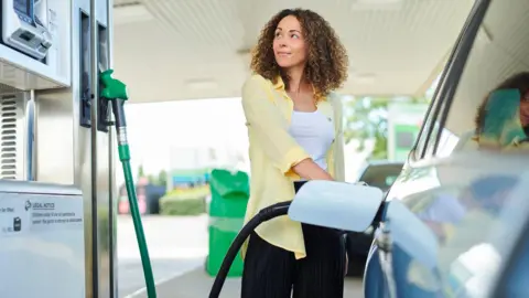 A woman wearing a white top and yellow shirt looks at a pump while filling up her car.