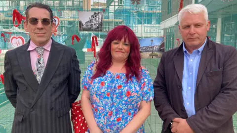 A man wearing a suit, stood alongside a women wearing a blue flowery dress, and another man in a suit - behind them is fencing protecting a large black and white derelict building. 