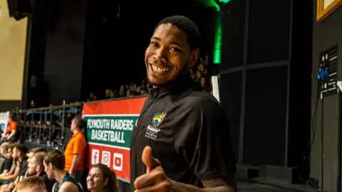 Kinu Rochford smiles widely and gives a thumbs up to the camera with a Plymouth Raiders Basketball banner held up behind him among a crowd.