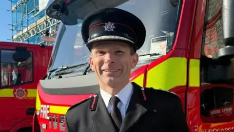 Jon Le Page smiles as he stands in front of two fire engines. He is wearing his uniform.