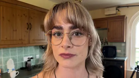 Georgina Cox with blonde hair, dark eyebrows and glasses - standing in a kitchen looking thoughtful with a slight smile.