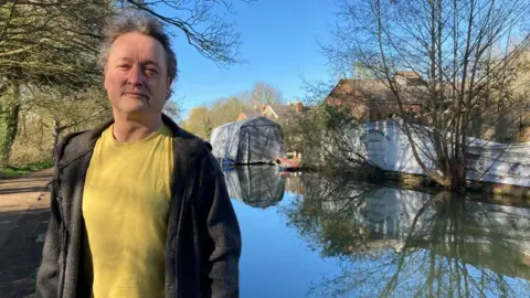A man in a yellow shirt is standing to the left of the shot. Behind him is a river and the site of a potential new boatyard.