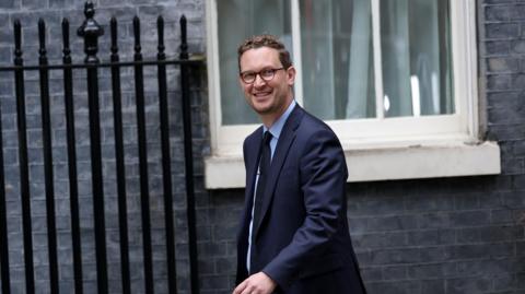 Darren Jones wearing a dark suit and dark, tie wearing glasses with short dark hair, smiling at the camera as he walks to Downing Street
