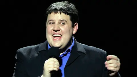 Jo Hale/Getty Images Peter Kay on stage. He is wearing a dark suit jacket and a purple shirt and is holding a microphone. He is bathed in a spotlight and set against a black backdrop.