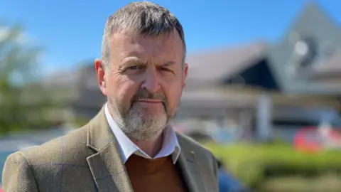 Dr Martin Rankin has short grey hair and a short grey bears. He is wearing a light brown blazer, with a white shirt under a brown jumper underneath. He is standing in front of the Noble's Hospital entrance which is blurry, and there are green bushes and trees behind him. It is a sunny day with cloudless blue skies.