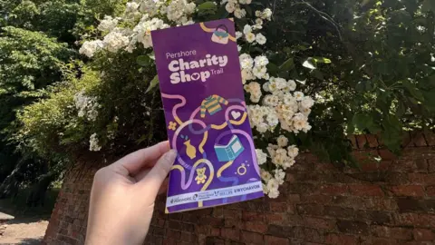 Wychavon District Council A hand holding a leaflet up in front of some white flowers bloming over a wall. The leaflet is purple and titled "Pershore Charity Shop Trail".