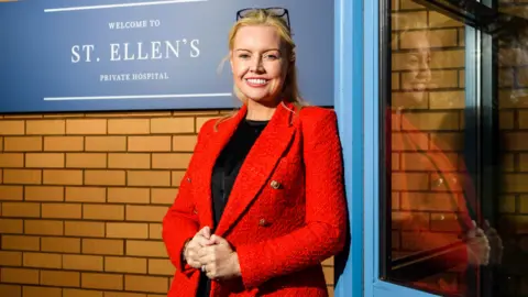 Gill Baird, who has blonde hair tied back, smiles at the camera while standing outside a brick building. She is wearing a red jacket over a black top. 