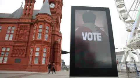 People walk near an election information sign reading "vote" near the Senedd (Welsh Parliament) building and the red Pierhead building in Cardiff Bay.