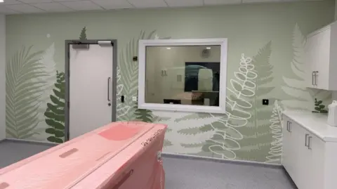 NHS A testing room at the new centre. There is a bed under a plastic sheet covering in a room with one wall covered in green wallpaper with a leaf pattern. There are white cabinets to the right of the room.