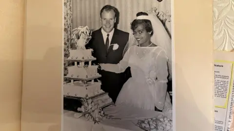A black and white wedding photo of Michael and May Tanner. They are smiling at the camera and cutting their wedding cake.