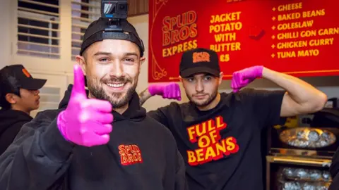 Jacob (left) and Harley Nelson in the kitchen in front of a red menu. Both are wearing black baseball hats and tops. Jacob is doing a thumbs up gesture while wearing a pink latex glove and Harley is in the background flexing his muscles